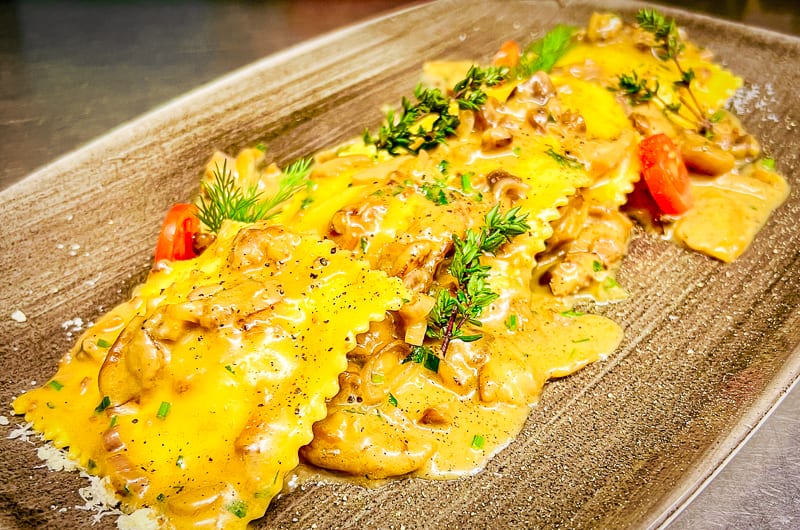 Rechteckige braune Platte mit gelben Ravioli überzogen mit Steinpilzsaoße, garniert mit Kräuterstängeln und roten Kirschtomaten.