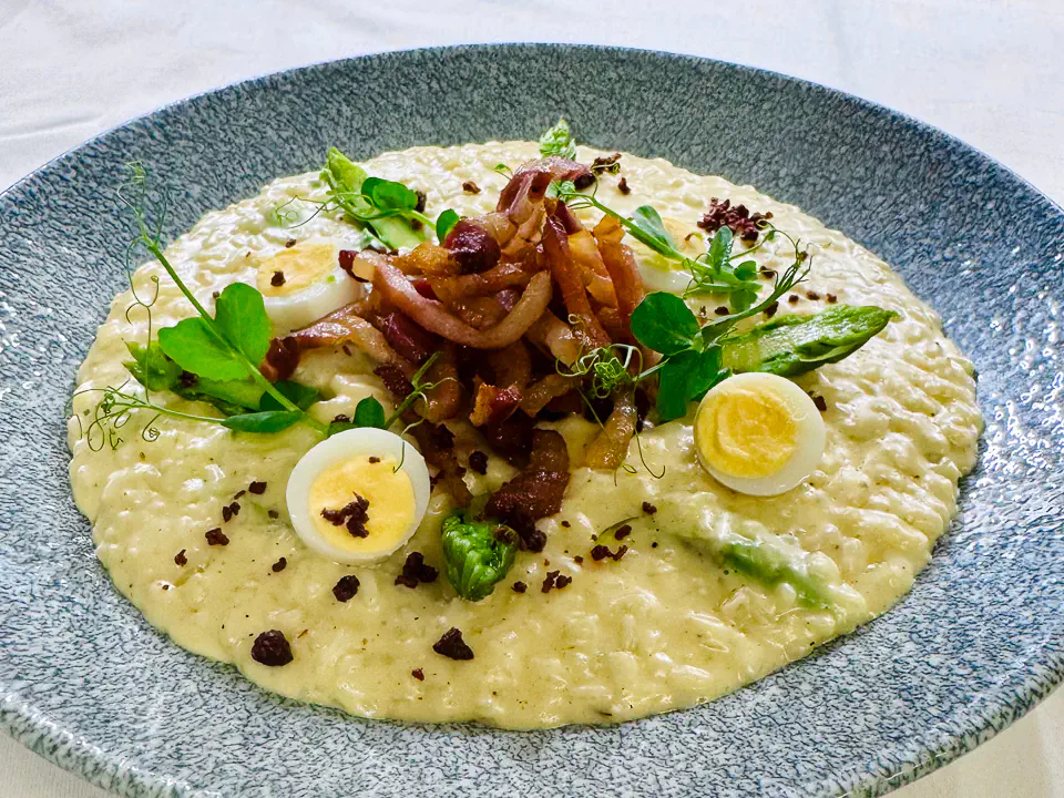 Ein cremiges Risotto-Gericht, angerichtet auf einem strukturierten, graublauen Teller. Das Risotto ist mit grünem Spargel, knusprigem Speck und halbierten Wachteleiern verfeinert. Frische Microgreens und knusprige Brösel runden die Dekoration ab.