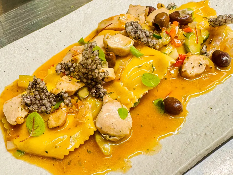 Spargel-Ravioli mit hellem Kaninchenfilet-Ragout und Taggiasca-Oliven, serviert in einer aromatischen Sauce. Dazu feine Gemüsestücke, knusprige Tapioka-Chips und frische Microgreens, angerichtet auf einem rechteckigen, hellen Teller.