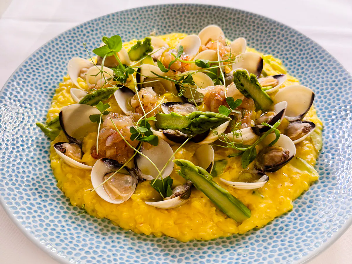 Mailänder Risotteo mit Venusmuscheln, grünem Spargel und violettem Garnelentatar, angerichtet auf einem hellblauen, strukturierten Teller und garniert mit frischen Microgreens.