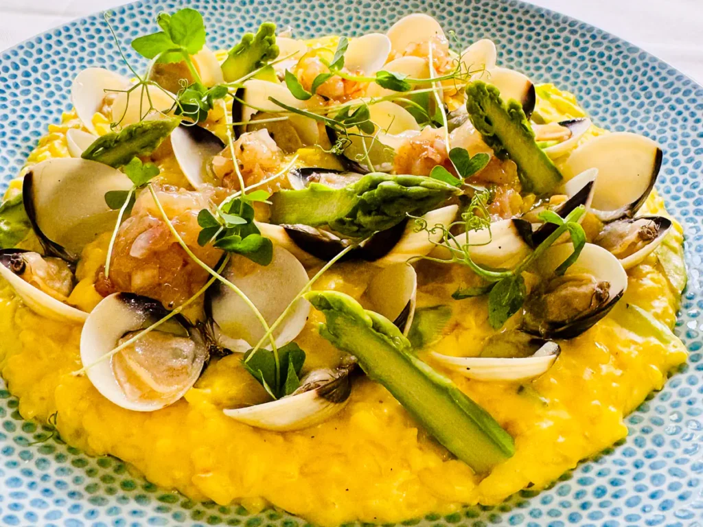 Mailänder Risotteo mit Venusmuscheln, grünem Spargel und violettem Garnelentatar, angerichtet auf einem hellblauen, strukturierten Teller und garniert mit frischen Microgreens.