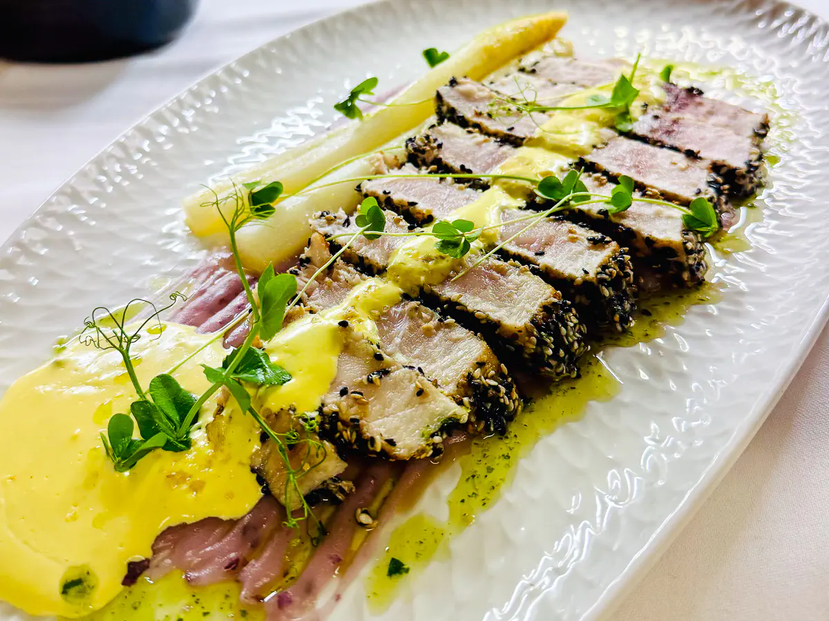In Sesamkruste gebratenes Schwertfisch-Tataki, angerichtet mit weissem Spargel, violetter Kartoffelcreme, gelber Hollandaise und grünem Kräuteröl, garniert mit frischen Microgreens auf einem ovalen, weissen strukturierten Teller.