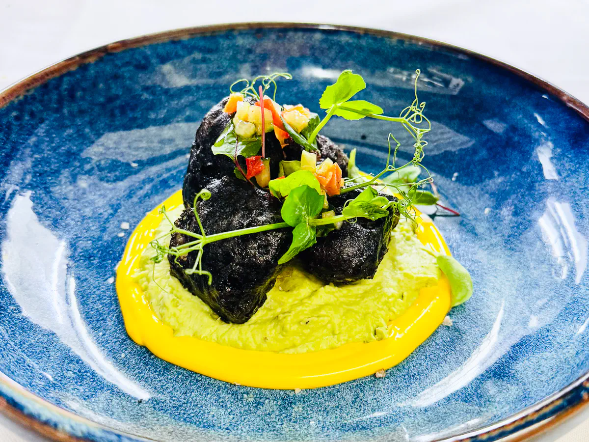 Zwei knusprige Würfel vom Zackenbarsch in schwarzer Sepiatempura auf einem Bett aus grüner Frühlings-Bohnenvelouté, umrahmt von leuchtend gelber Yuzu-Mayonnaise und garniert mit fein geschnittenem Gemüse sowie frischen Microgreens, serviert auf einem tiefen, blauen Teller.