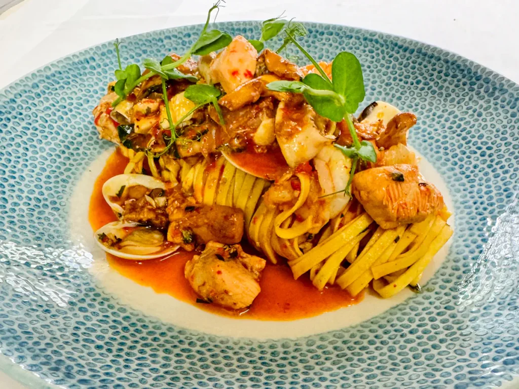 Frische Tagliolini auf einem blau-weiss gemusterten Teller, serviert mit Meeresfrüchten, Muscheln, Waldpfifferlingen und Stückchen vom Fang des Tages, überzogen mit einer roten Paprikasauce und garniert mit frischen Kräutern.