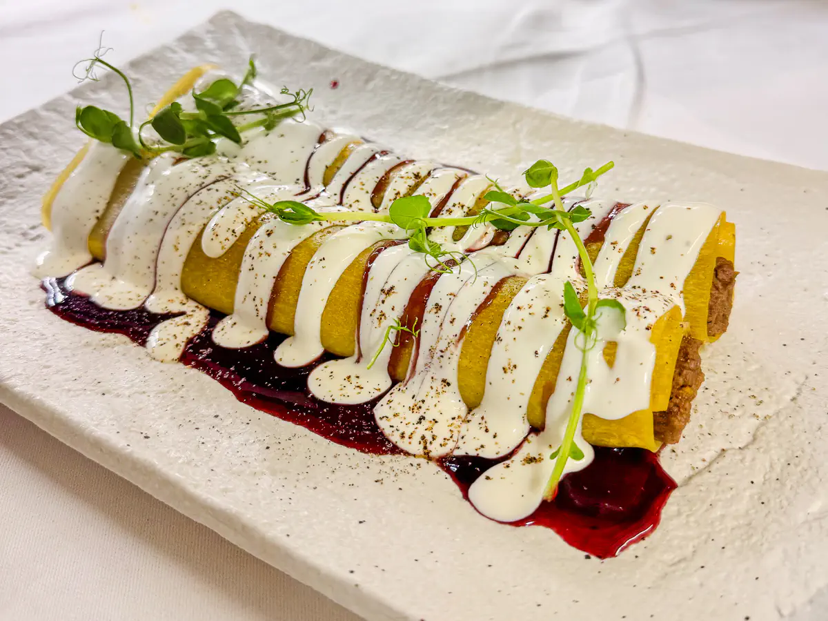 Offene Lasagne-Röllchen auf weissem Teller, gefüllt mit Rinderfilet, angerichtet auf einer dunklen Portweinreduktion, überzogen mit heller Zitronen-Ziegenrobiola-Creme und garniert mit frischen Kräutern.