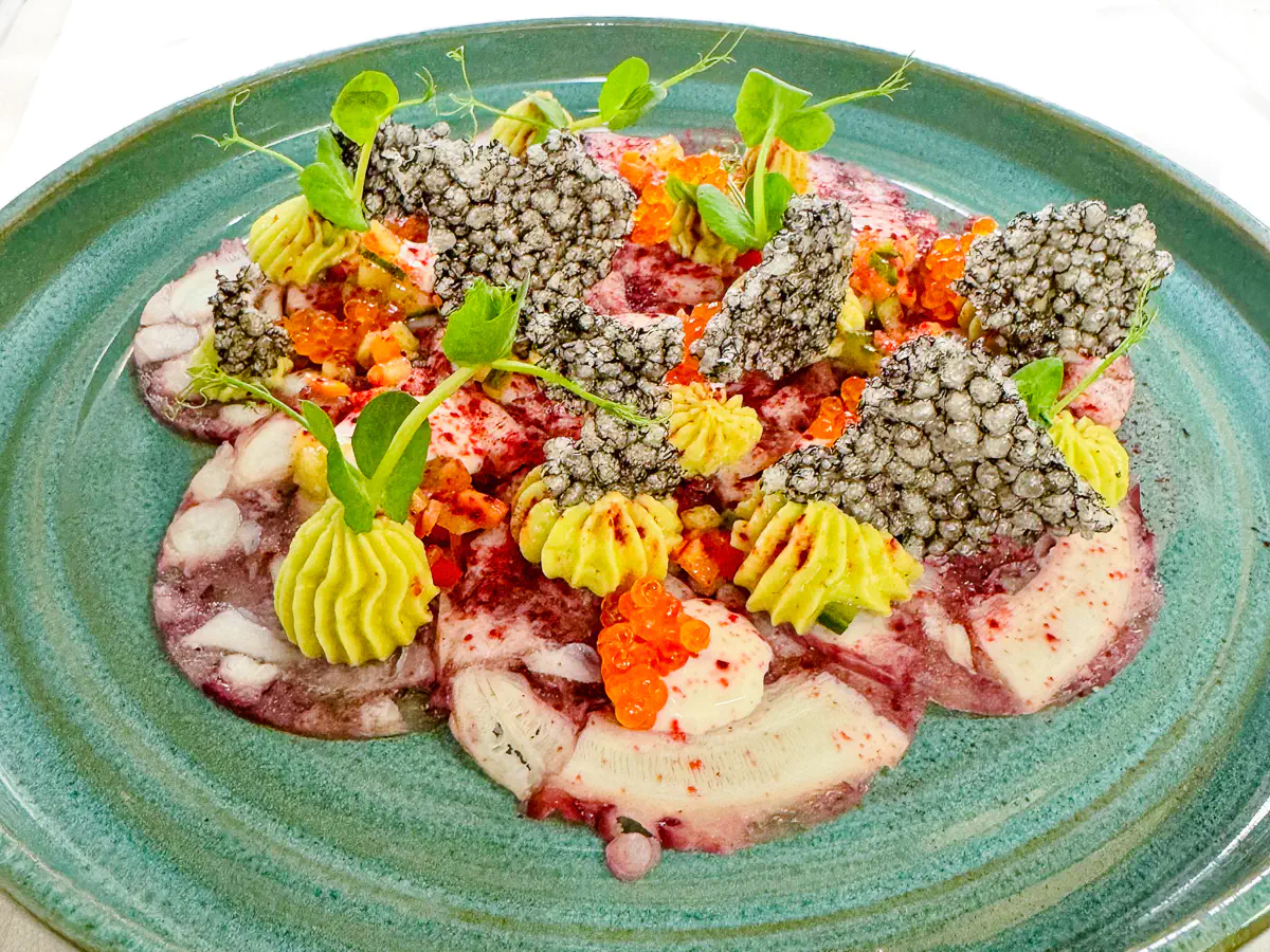 Kunstvoll angerichtetes Carpaccio vom Oktopus auf einem grünen Teller, garniert mit grünem Avocadomousse, Burrataschaum, orangefarbenem Zitronenkaviar, knusprigen schwarzen Tapioka-Chips, roten Pulverakzenten von Rote Bete und frischen Sprossen.