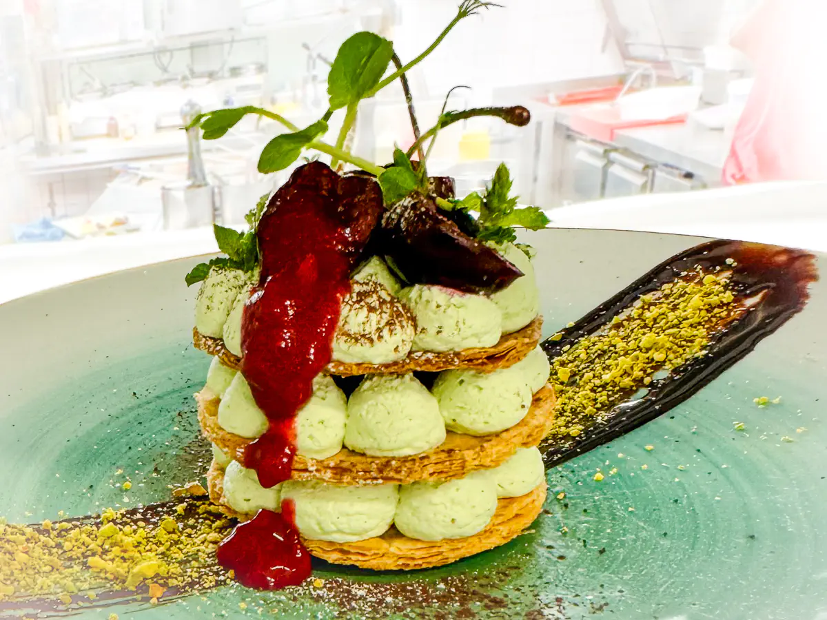 Auf einem grünen Teller ist ein hohes Blätterteig-Dessert angerichtet, das aus drei Schichten knusprigem Blätterteig besteht, dazwischen Tupfer von weisser Schokoladen-Matcha-Chantilly. Obenauf liegt ein Kirsch-Tatar, das leicht über die Seiten läuft, garniert mit frischen Kräutern. Der Teller ist mit einem Pinselstrich aus dunkler Zartbitterschokolade und gehackten Pistazien verziert, dazu etwas Pistazien-Creme anglaise.