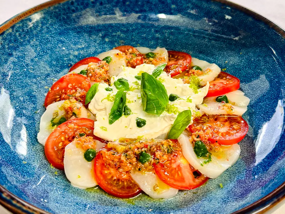 Auf einem tiefen blauen Teller ist eine moderne Caprese di mare angerichtet. In einem Kreis wechseln sich dünne Scheiben gebratener Jakobsmuscheln und marinierter süsser Tomaten ab. In der Mitte liegt ein cremiger Klecks frischer Stracciatella, garniert mit Basilikumblättern. Feine grüne Tropfen und ein Hauch Vanille-Limette-Olivenöl runden das farbenfrohe Gericht ab.