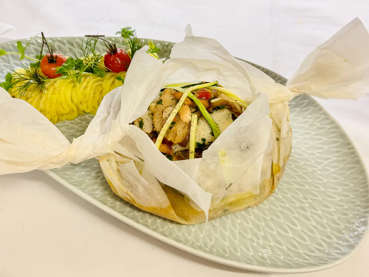Auf einem hellen, länglichen Teller liegt ein im Pergamentpapier gegartes Fischpäckchen, das an den Enden kunstvoll zugebunden ist. Daraus schaut ein Mix aus Fischstücken, Pfifferlingen, Steinpilzen, feinen Zucchinistreifen und Kräutern hervor, leicht mit Pfeffer bestreut. Daneben befindet sich ein dekorativ aufgespritztes Kartoffelpüree, beträufelt mit Olivenöl und garniert mit frischem Dill und zwei kleinen gebratenen Cherry-Tomaten. Im Fischgericht ist eine Safran-Zitronen-Sauce angedeutet.