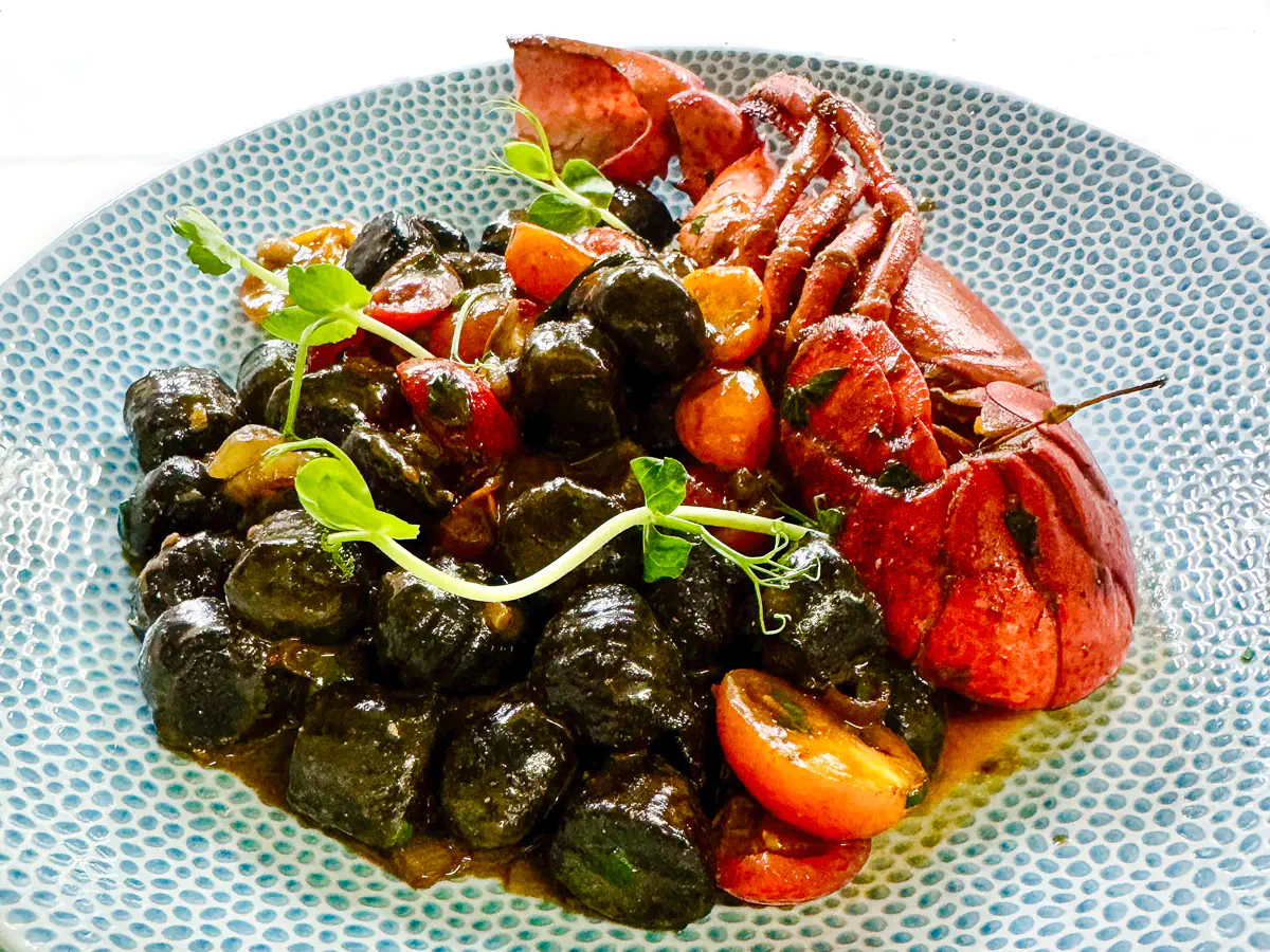 Auf einem hellblau gemusterten Teller liegen von Hand gefertigte schwarze Gnocchi mit Sepiatinte, vermengt mit einer aromatischen Hummerbisque. Dazu sind ein gebratener Hummerschwanz, halbierte rote und gelbe confierte Datteltomaten sowie einige zarte grüne Kräuter angerichtet. Ein Hauch Brandy rundet das Gericht ab.
