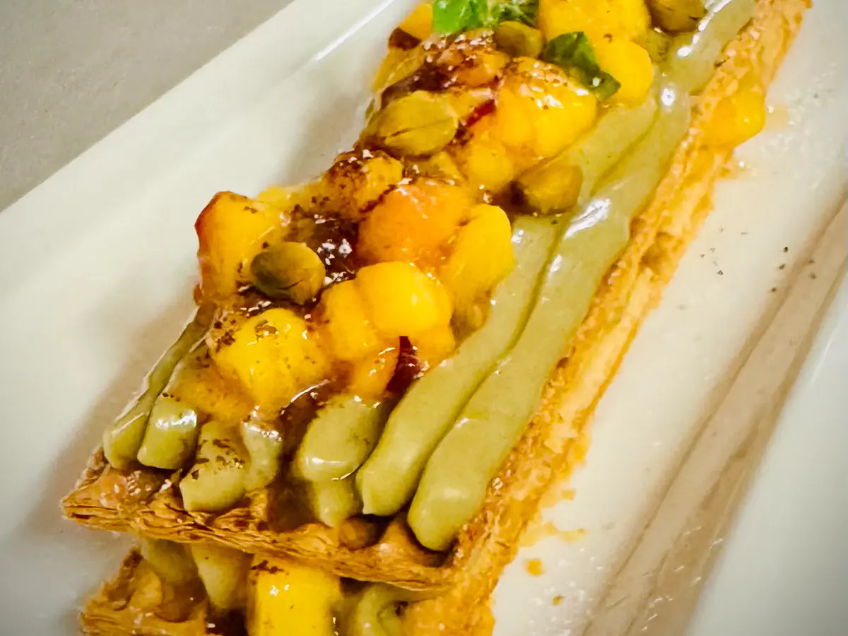 Auf einem rechteckigen weissen Teller liegt eine karamellisierte Millefeuille aus knusprigem Blätterteig, gefüllt und garniert mit grüner Bourbon-Vanille-Limetten-Crème. Obenauf befinden sich gewürfelte Stücke von frischer Mango und in Rosmarin-Osmose marinierter Pfirsich, dekoriert mit ein paar Pistazien und einem Hauch frischer Kräuter.