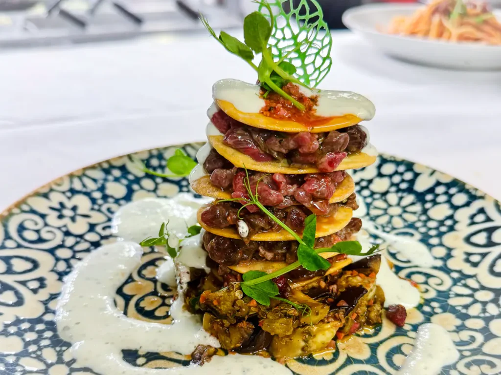 Auf einem kunstvoll gemusterten Teller ist eine hohe Millefoglie aus dünnen Scheiben “Lingua di suocera”-Blätterteig geschichtet, gefüllt mit gewürztem Lamm-Tartar. Dazwischen befindet sich eine geräucherte Auberginencreme, die zusammen mit der hellen Joghurt-Limetten-Minzsauce über die Ränder läuft. Frische Kräuter und ein filigranes grünes Dekorgitter krönen das Gericht.