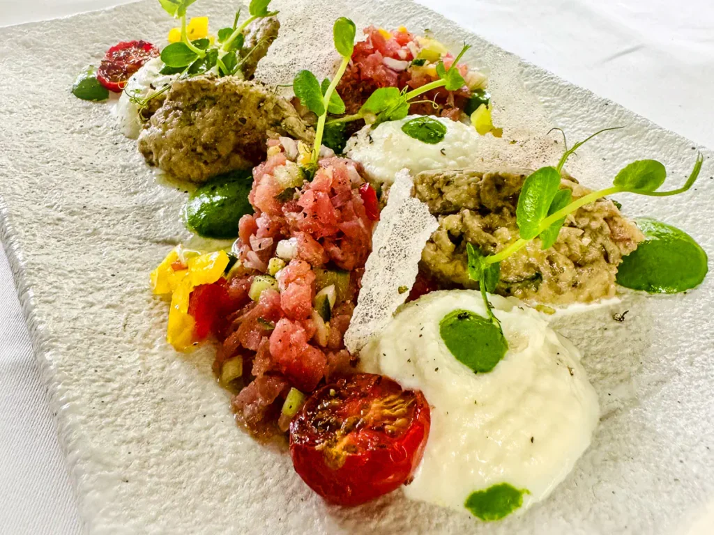 Auf einem rechteckigen, hell strukturierten Teller ist eine moderne Variante der Parmigiana di Melanzane angerichtet. Es finden sich Häufchen von gegarter Aubergine der Sorte “Violetta di Firenze”, dazu fein gewürfeltes Tatar vom roten Thunfisch, confierte Datteltomaten, gelbe und rote Paprikastückchen sowie Kleckse einer leuchtend grünen Basilikumcreme. Ergänzt wird das Ganze durch weissen, luftigen Büffelmozzarella-Schaum und zarte grüne Kräuter.