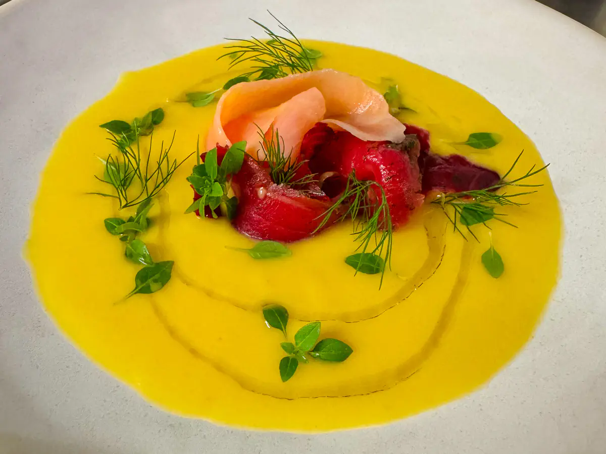 Auf einem weissen Teller ist eine leuchtend gelbe Pfirsich-Mango-Gazpacho angerichtet. In der Mitte liegt dekorativ gerolltes Carpaccio vom geräucherten Lachs, daneben kleine Stücke von roter Beete oder ähnlich farbigem Gemüse. Die Suppe ist mit frischen grünen Sprossen, Dill und feinen Blättchen garniert, darüber sind zarte Kreise Olivenöl gezogen. Fein geriebene Limettenschale rundet das Arrangement ab.