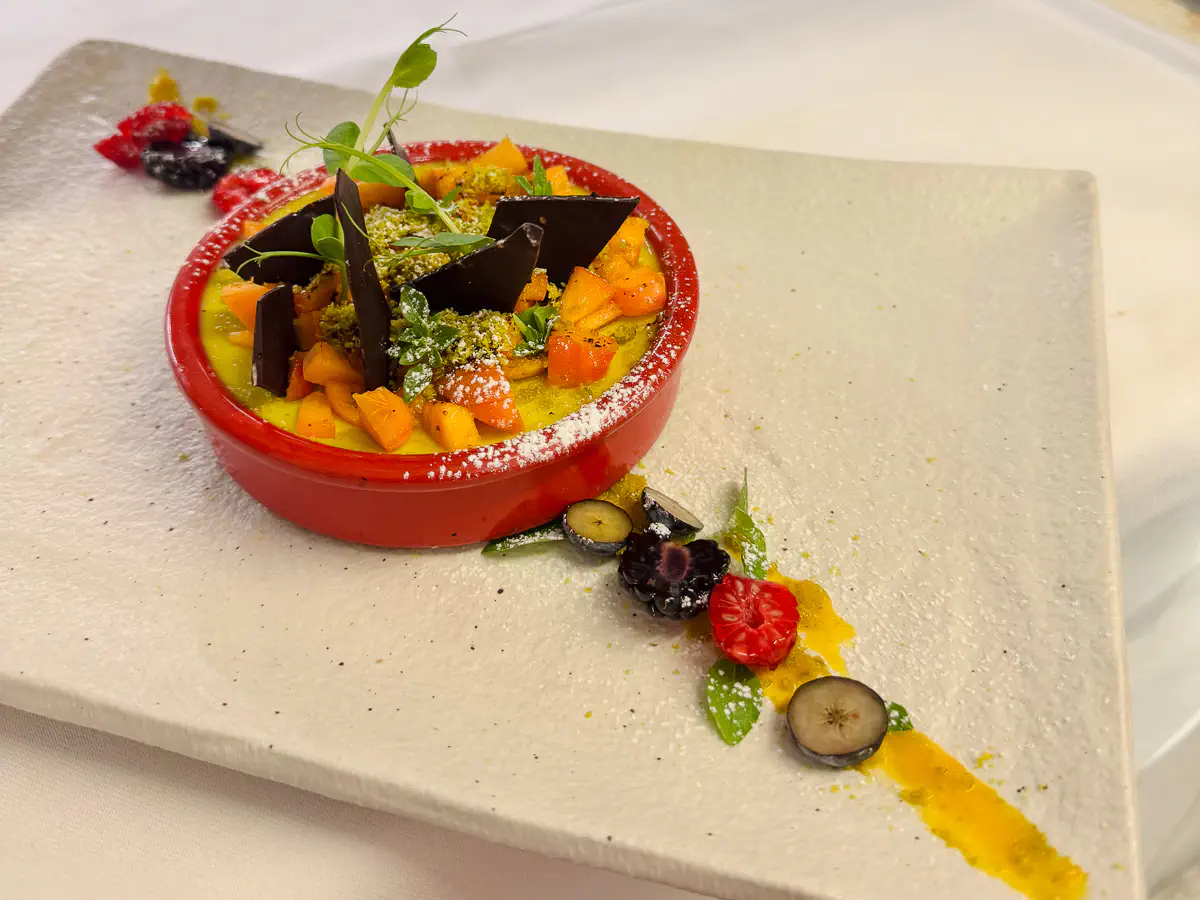 Auf einem hellen, eckigen Teller steht eine rote Auflaufform, gefüllt mit einer Pistazien-Crème-Brûlée. Die Oberfläche ist mit gewürfelten Aprikosenstücken, dunklen Schokoladensplittern, grünem Basilikum und Puderzucker garniert. Auf dem Teller verlaufen ein gelber Fruchtspiegel sowie frische Beeren wie Himbeeren, Brombeeren und halbierte Heidelbeeren, ergänzt durch grüne Blätter und etwas Puderzucker.