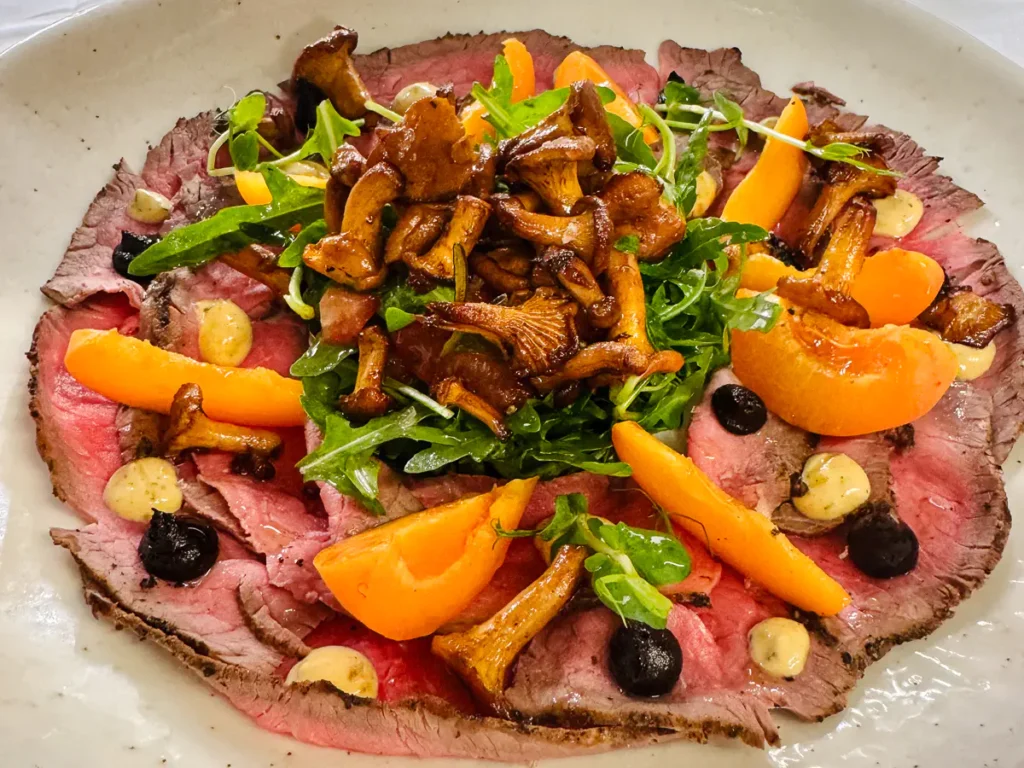Auf einem grossen, hellen Teller ist dünn aufgeschnittenes, rosa gegartes Roastbeef vom Piemonteser Jung-Rind angerichtet. Darauf liegen geröstete Pfifferlinge, Wildrucola und Spalten von frischen Aprikosen. Tupfer einer Aprikosen-Senf-Creme und schwarzer Knoblauch sind dekorativ verteilt. Geröstete Mandeln und ein Hauch geräuchertes Maldon-Salz runden das Gericht ab.