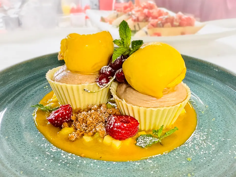 Auf einem türkisfarbenen Teller stehen zwei weisse Schokoladenkörbchen, gefüllt mit cremigem Erdnussmousse. Obenauf jeweils eine Kugel leuchtend gelbes Mangosorbet. Das Dessert ist umgeben von einem Mango-Fruchtspiegel, frischen Himbeeren, Minze und knusprigem Meersalz-Crunch, der dem Gericht Textur verleiht. Leicht mit Puderzucker bestäubt.
