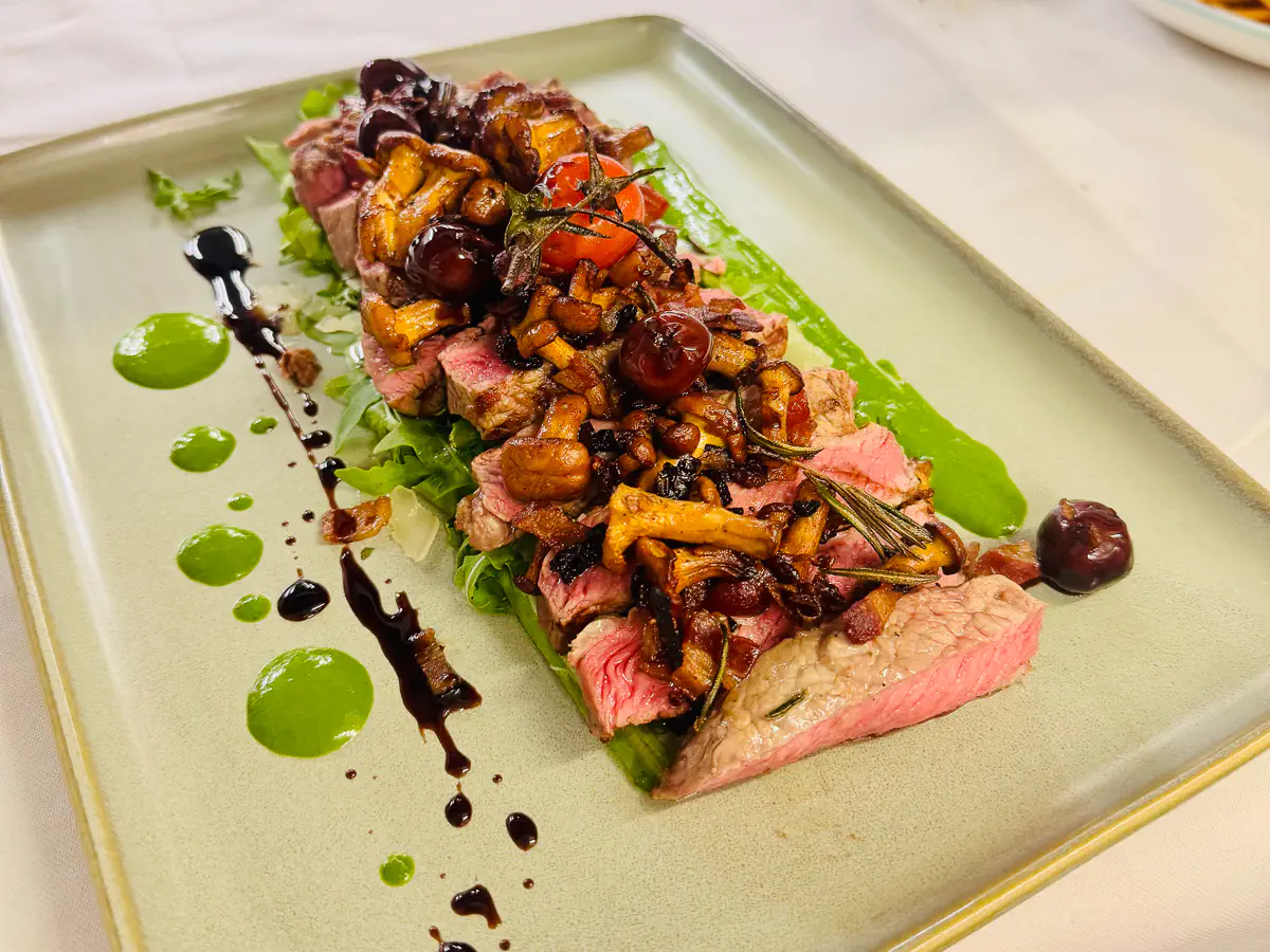 Auf einem rechteckigen, hellgrünen Teller liegt eine Tagliata vom rosa gebratenen Piemonteser-Fassona-Controfilet, angerichtet auf einem Bett aus frischem Rucola. Das Fleisch ist belegt mit knusprigen Stücken Tiroler Speck, gebratenen Pfifferlingen und Grana Padano. Darüber sind dunkle Balsamico-Kirschen verteilt. Am Tellerrand befinden sich Tupfer von grüner Sauce und dunklem Balsamico-Reduktion.