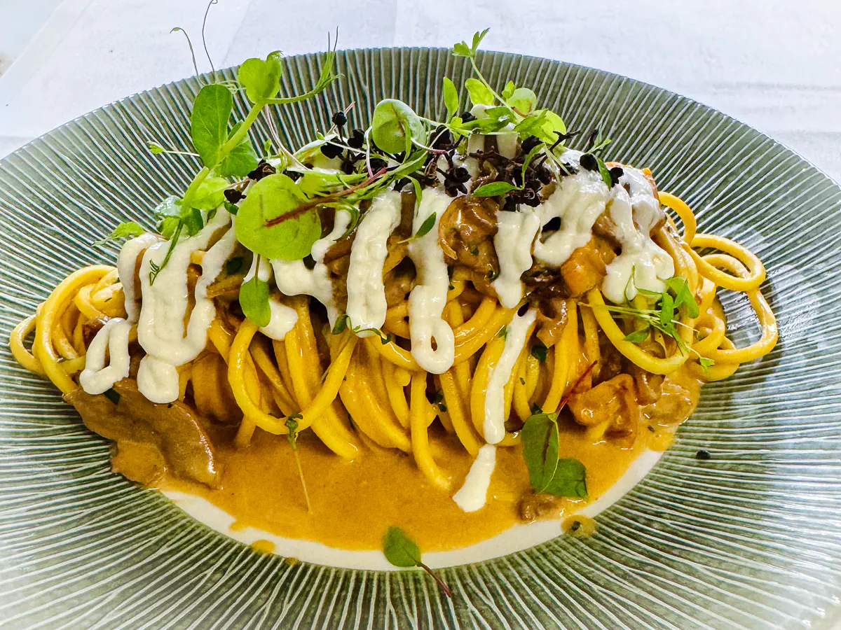 Auf einem grün gerillten Teller liegt ein Nest aus Chitarra-Spaghetti, umhüllt von einer cremigen Sauce mit karamellisierten Aprikosen, verfeinert mit Kastanienhonig. Obenauf zart rosa gebratene Stücke von Entenbrust, darüber Fäden von geschmolzenem Pecorino Sardo DOP. Frische Kräuter und feine Sprossen runden das Gericht optisch und aromatisch ab.