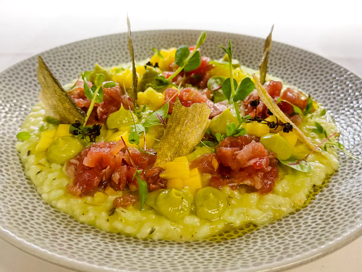 In einem tiefen, graugemusterten Teller ist ein cremiges Limetten-Risotto aus gereiftem Carnaroli-Reis angerichtet. Obenauf liegt fein gewürfelter roher roter Thunfisch, begleitet von leuchtend gelben Mangowürfeln und kleinen Avocadocreme-Tupfern. Knusprige Chips und zarte Kräuter sorgen für Struktur und Frische, verfeinert mit aromatischem Timut-Pfeffer.