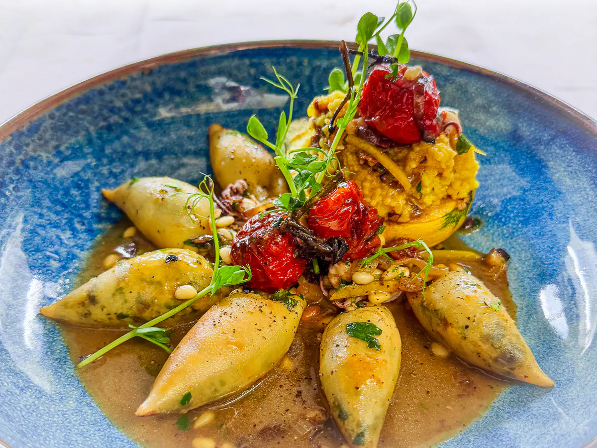 Auf einem tiefblauen Teller sind zarte Calamaretti angerichtet, gefüllt mit einer mediterranen Kräuter- oder Fischfarce. Die Tintenfische liegen in einer aromatischen Jus mit Pinienkernen und frischen Kräutern. Dazu eine gefüllte runde Zucchini mit Couscous und gebratenem Gemüse, getoppt mit konfierten Kirschtomaten und dekorativem Grün.