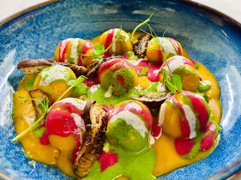 In einem tiefen blauen Teller liegen grosse, gefüllte Gnocchoni auf einem leuchtend orangefarbenen Kürbisvelouté. Die Gnocchi sind mit Steinpilzen gefüllt und mit bunten Streifen aus geschmolzenem Gorgonzola dolce, grüner Kräutersauce und roter Beete-Creme verziert. Dazwischen gebratene Steinpilze, knuspriger Kastanien-Crumble und karamellisierte Pekannüsse, garniert mit frischen Kräutern.