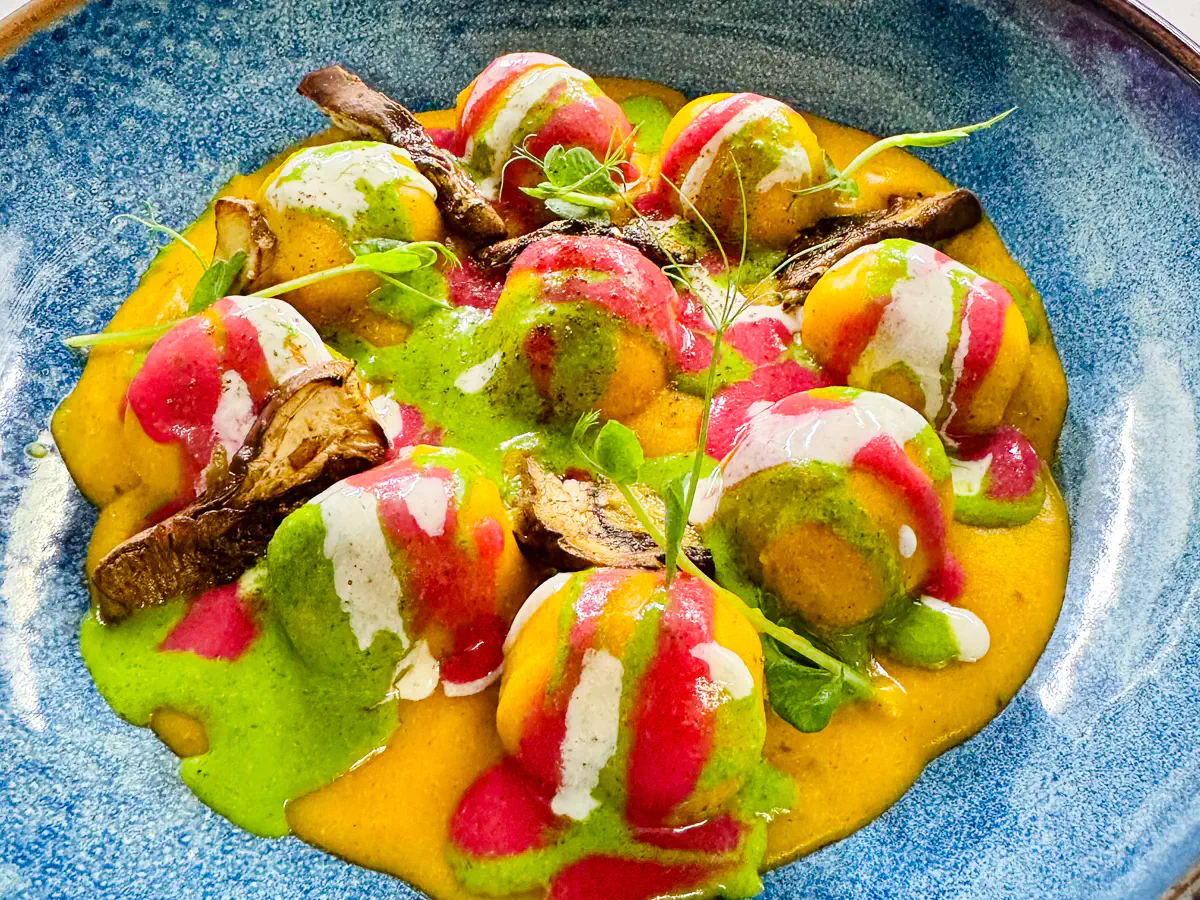 In einem tiefen blauen Teller liegen grosse, gefüllte Gnocchoni auf einem leuchtend orangefarbenen Kürbisvelouté. Die Gnocchi sind mit Steinpilzen gefüllt und mit bunten Streifen aus geschmolzenem Gorgonzola dolce, grüner Kräutersauce und roter Beete-Creme verziert. Dazwischen gebratene Steinpilze, knuspriger Kastanien-Crumble und karamellisierte Pekannüsse, garniert mit frischen Kräutern.