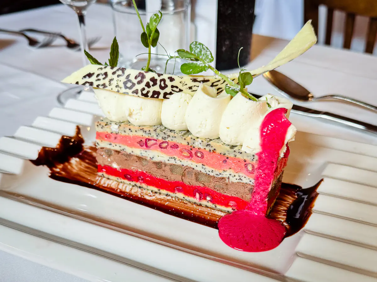 Ein kunstvoll geschichtetes Dessert aus Moelleux-Biscuits mit Mohn, gefüllt mit Lagen von karamellisierter Birne, Zartbitterschokoladen-Mousse und leuchtend roter Granatapfel-Crème-Anglaise. Das Dessert ist mit Tupfen heller Creme, einer dekorativen weissen Schokoladenscheibe im Animal-Print und frischen Kräutern garniert. Auf dem Teller verlaufen zusätzlich Schokoladensauce und ein Fruchtspiegel.