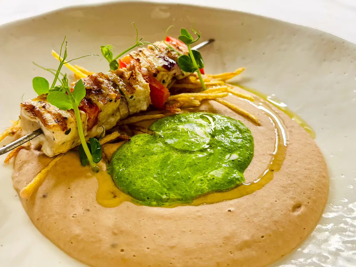Auf einem hellen Teller liegt ein Spiess mit zart gegrillten Würfeln vom Schwertfisch, abwechselnd mit roten Paprikastücken. Der Spiess ruht auf einem cremigen Cannellini-Hummus, in dessen Mitte eine leuchtend grüne mediterrane Salsa Verde angerichtet ist. Feine Streifen knuspriger Kartoffeln und frische Kräuter runden das Gericht ab, dezent bestreut mit Pistaziengranella.