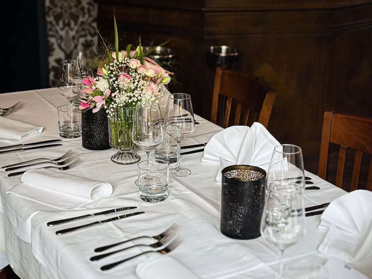 Elegant gedeckter Tisch mit weisser Tischdecke, weissen Stoffservietten in Fächerform und vollständigem Besteck für mehrere Gänge. In der Mitte ein Blumenarrangement aus rosa und weissen Blüten in einem grünen Glasgefäss, daneben schwarze Windlichter mit Struktur. Gläser für Wasser, Wein und Sekt sind sorgfältig platziert, umgeben von Holzstühlen in stilvollem Ambiente.