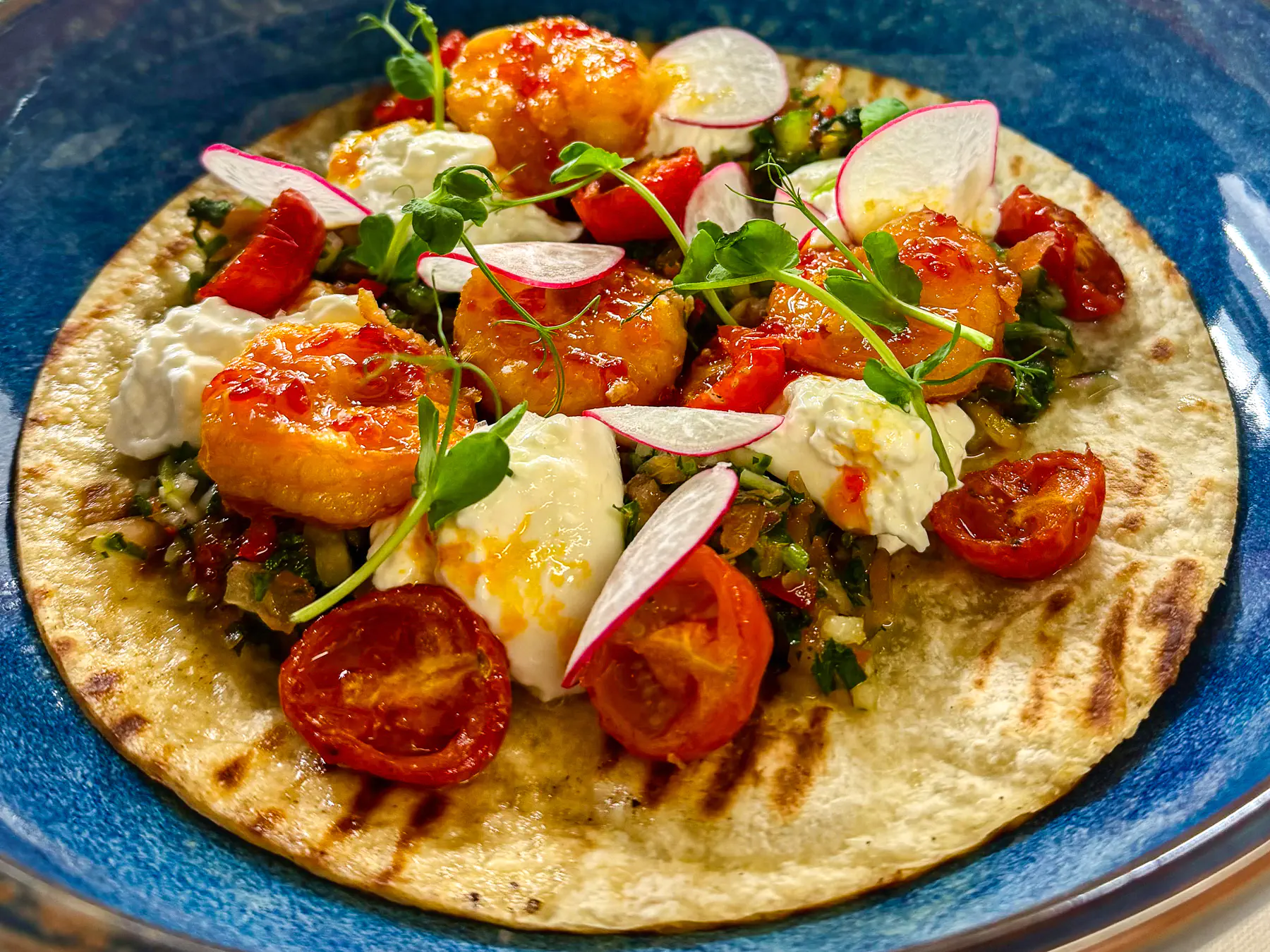 Auf einem blauen Teller angerichtete goldbraune Garnelen auf einer leicht gerösteten Fladenbasis, verfeinert mit weisser Creme, roten Tomaten, feinen Radieschenscheiben und frischen Microgreens.