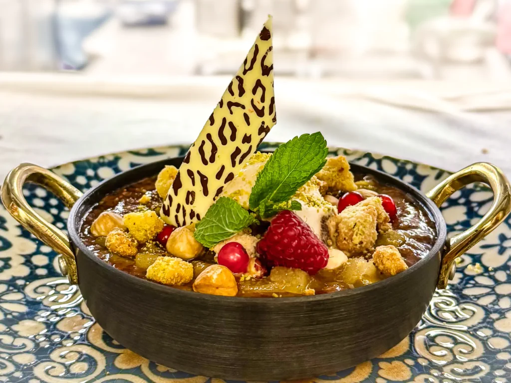 In einer kleinen schwarzen Servierpfanne mit goldenen Henkeln angerichtetes Dessert mit cremiger Brûlée-Oberfläche, garniert mit Beeren, Haselnüssen, Crumble, Minze und einem Stück weisser Schokolade.
