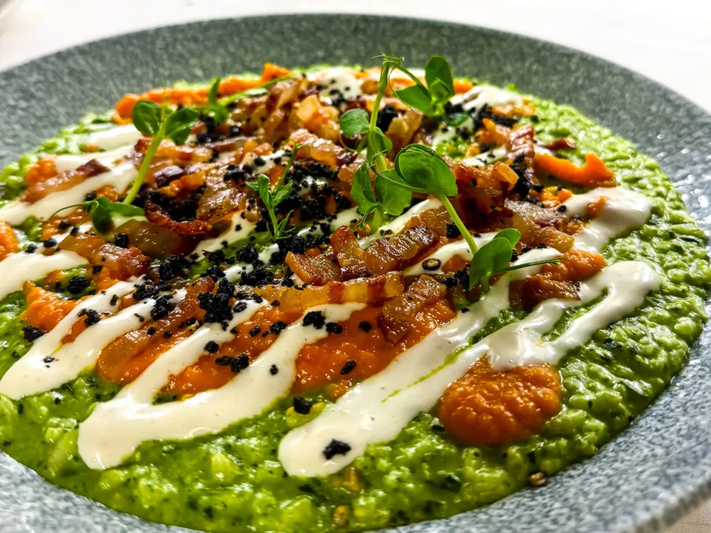 Auf einem grauen Teller serviertes leuchtend grünes Risotto mit orangefarbener Kürbiscreme, weissen Pecorino-Streifen, knusprigem Speck und frischen grünen Microgreens als Garnitur.
