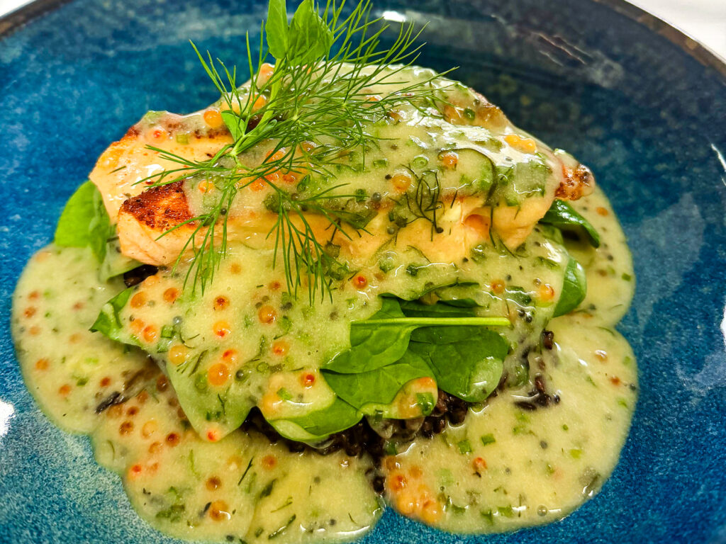 Auf einem tiefblauen Teller angerichtetes Lachsfilet auf dunklem Venere-Reis und frischem Spinat, überzogen mit heller, cremiger Sauce mit Senfkörnern und zartem Dill als Garnitur.