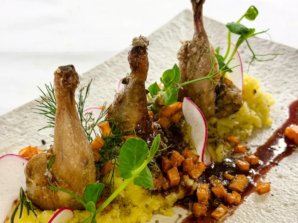 Auf einem strukturierten, hellen Teller angerichtete goldbraune Wachtelkeulen auf cremigem Kartoffelpüree, umgeben von leuchtend orangefarbenen Kürbiswürfeln, roten Radieschenscheiben, Kräutern und dunkler Sauce.