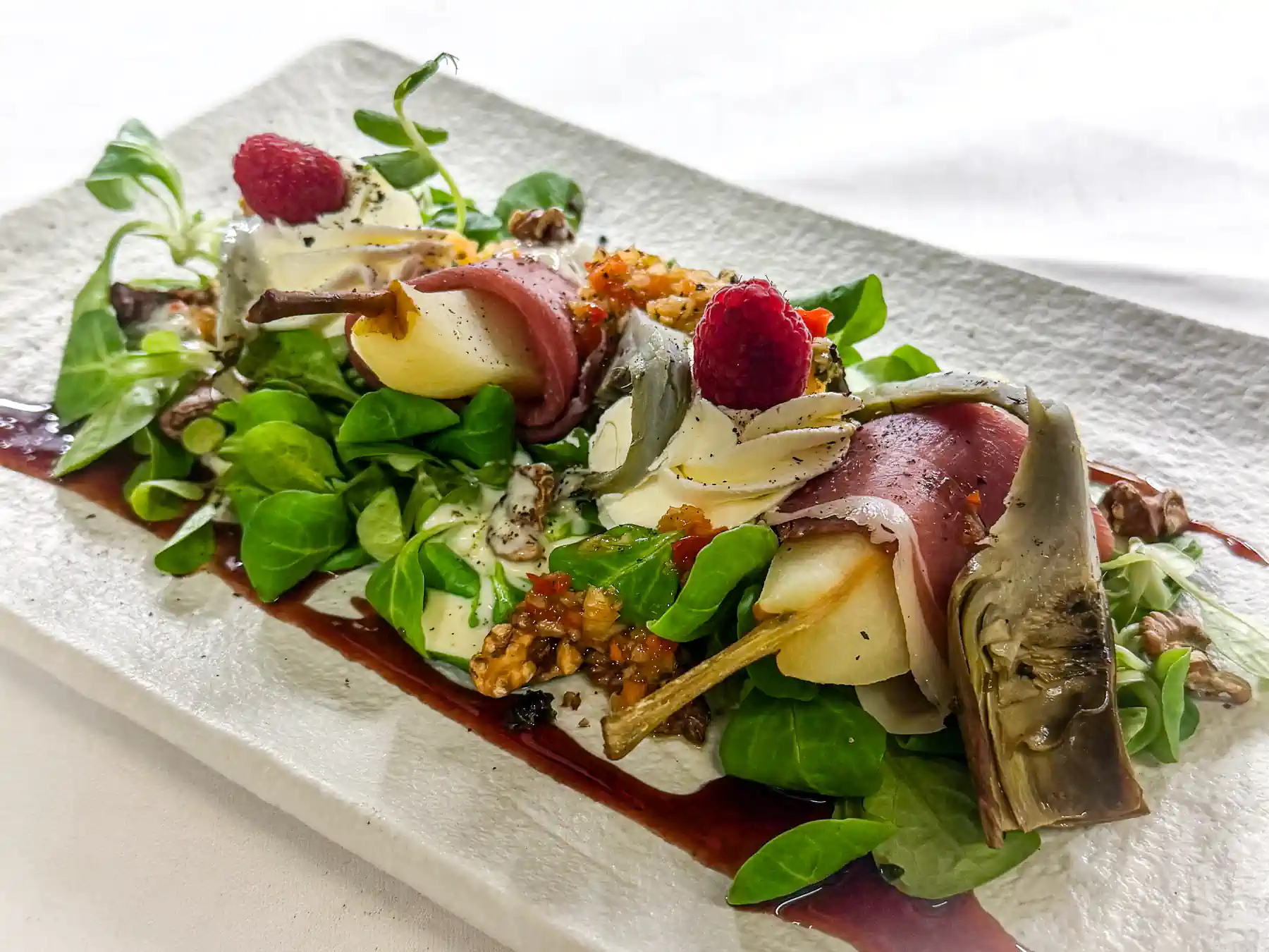 Auf einem rechteckigen, hellen Teller angerichteter Salat mit zarten Artischocken, Birnenspalten, Parmaschinkenröllchen, weisser Käsemousse, frischen Himbeeren, grünen Blättern und goldbraunen Walnüssen.