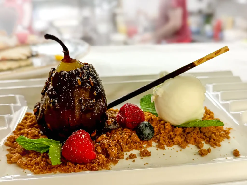 Auf einem rechteckigen, weißen Teller liegt eine dunkle, glänzende Schokoladenbirne auf goldenem Mandelcrumble, begleitet von einer runden Kugel Vanilleeis, frischen Beeren und einem feinen Schokostick – dekoriert mit Minzblättern.