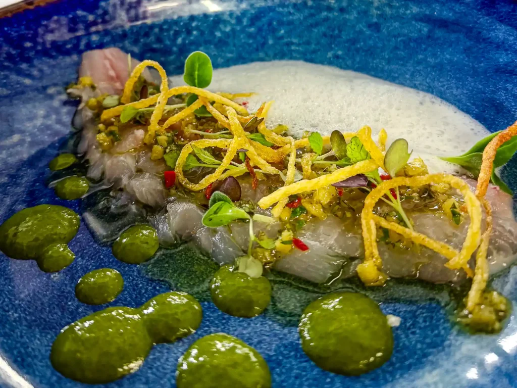 Auf einem tiefblauen Teller liegen zart-transparente Scheiben der marinierten Dorade, leicht glänzend und von feinen Kräutern durchzogen. Darüber verteilen sich knusprige, goldene Fäden und kleine grüne Blätter. Rundherum setzen intensiv grüne Tupfer und ein luftiger weißer Schaum frische, lebendige Akzente.