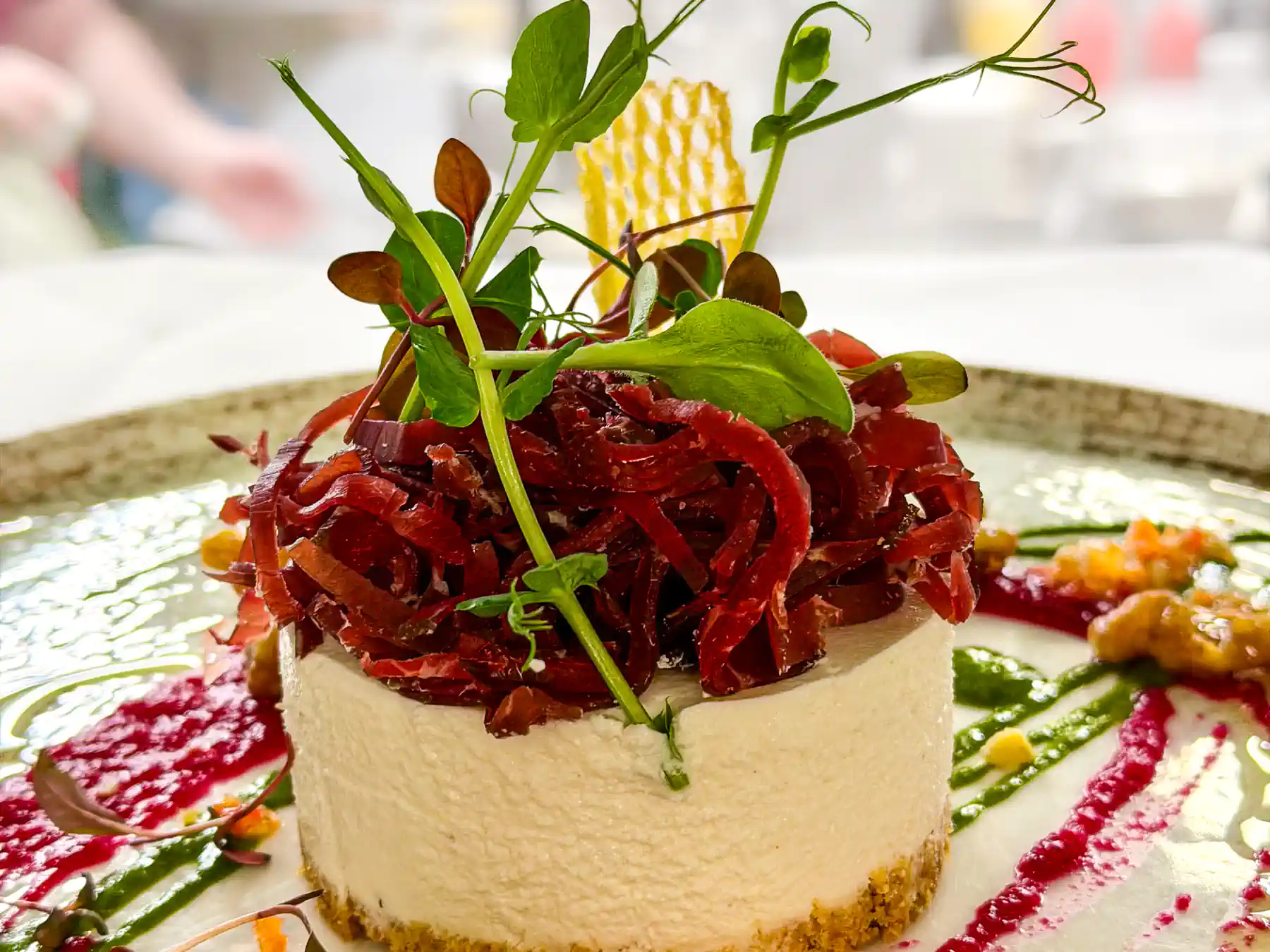 Auf einem rustikalen Keramikteller steht ein runder, heller Cheesecake aus Ziegenkäsemousse auf knusprigem Boden. Obenauf türmt sich ein farbintensiver Berg aus fein geschnittenem Bresaola, durchzogen von frischen grünen Blättern. Um den Cheesecake ziehen sich kräftige Pinselstriche in Rot und Grün sowie kleine Tupfer und Crunch-Elemente, die das Gericht lebendig wirken lassen.