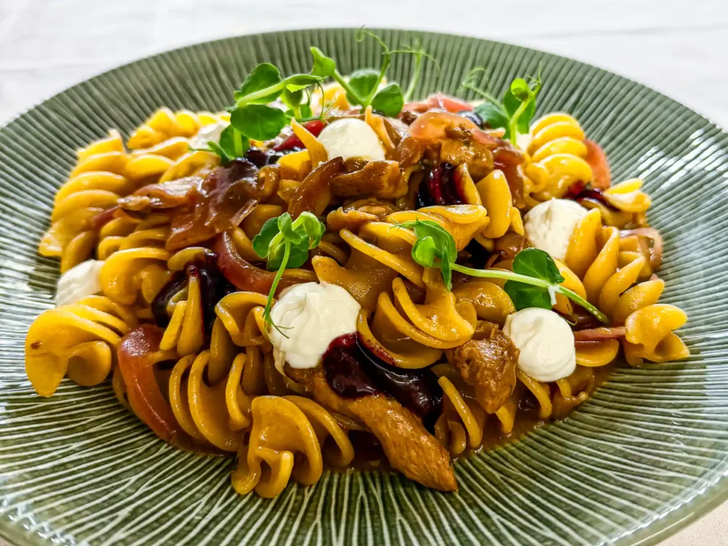 Auf einem grünen Teller angerichtete goldgelbe Fusillone-Pasta mit Stücken von Fleisch und Pilzen, verfeinert mit dunkler Sauce, roten Zwiebeln, weissen Cremetupfern und frischen Microgreens.