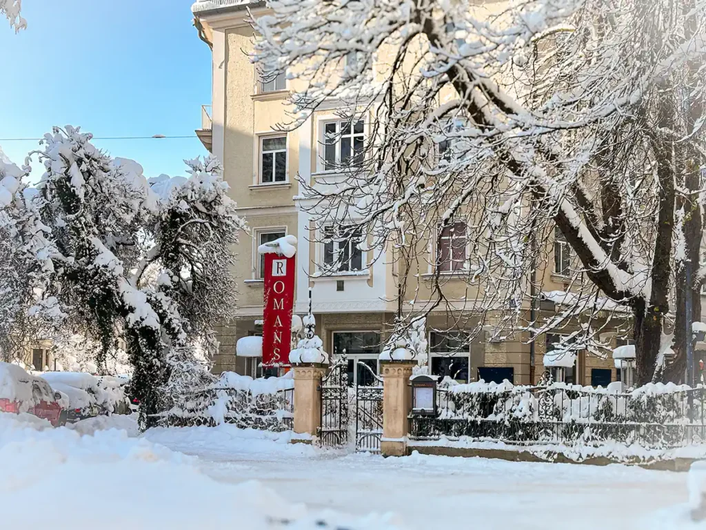 Winterliche Strassenszene mit dem Ristorante ROMANS im Hintergrund, vor dem eine rote Fahne mit der weissen Aufschrift "ROMANS" steht. Die Bäume und Sträucher im Vordergrund sind dicht mit Schnee bedeckt, ebenso wie der Boden, die Autos und der Zaun. Klare, blaue Himmelsfarbe und helles Tageslicht verstärken die kalte, ruhige Winteratmosphäre.