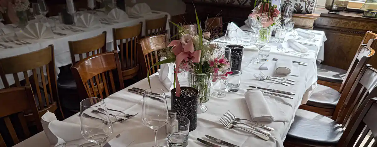 Feierlich gedeckter Tisch in einem Restaurant mit weisser Tischdecke, Stoffservietten, Gläsern, Besteck und schwarzen Windlichtern. In der Mitte des Tisches befinden sich Blumenarrangements mit rosa und weissen Blüten in Glasvasen. Die Tische sind von klassischen Holzstühlen umgeben und im Hintergrund sind weitere ähnlich gedeckte Tische sichtbar.
