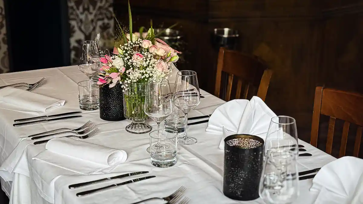 Eleganter, festlich gedeckter Tisch mit weisser Tischdecke, Stoffservietten, Besteck, Wassergläsern und Weingläsern. In der Mitte steht ein Blumenarrangement mit rosa und weissen Blüten in einer Glasvase sowie schwarze Windlichter. Die Holzstühle und die dunkle Wandverkleidung im Hintergrund verleihen dem Raum eine klassische Atmosphäre.