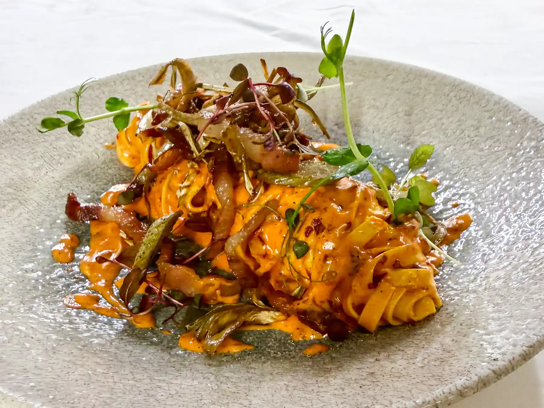 Auf einem großen, hellen Steinteller liegen Tagliatelle in einer leuchtend roten, cremigen Sauce, durchzogen von knusprigem Guanciale und gebratenen Artischocken. Feine Microgreens setzen frische grüne Akzente über dem warmen Pastaberg.
