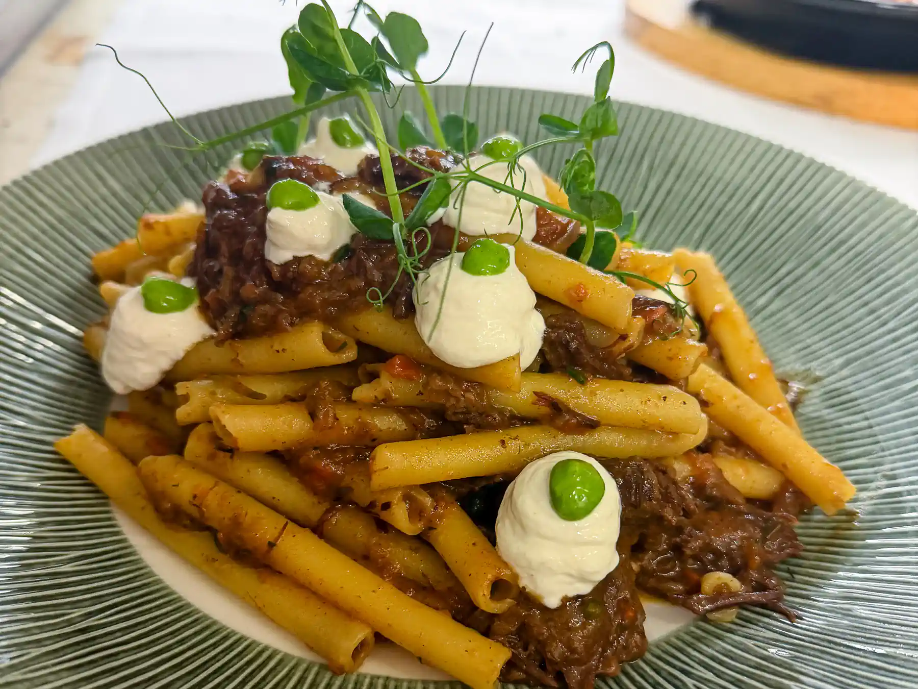 Auf einem grünen, gerillten Teller liegt ein üppiger Berg goldgelber Ziti, durchzogen von dunklem, glänzendem Schmorfleisch. Darüber sitzen kleine weiße Cremetupfen, jeweils mit einem runden, leuchtend grünen Akzent. Zarte Microgreens ragen wie feine Triebe nach oben und geben dem warmen, erdigen Farbenspiel eine frische, lebendige Note.