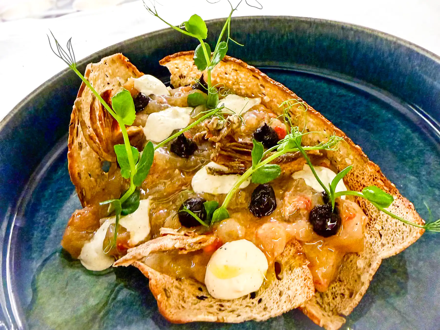 Auf einem tiefblauen Teller liegen knusprig geröstete Brotscheiben, locker übereinandergelegt. Darauf verteilt sich eine glänzende, fein strukturierte Masse in warmen Rosa- und Beigetönen, durchzogen von cremigen, hellen Tupfern. Dunkle Punkte setzen Akzente, während frische grüne Triebe und zarte Kräuter dem Gericht Höhe, Leichtigkeit und einen klar mediterranen Charakter verleihen.