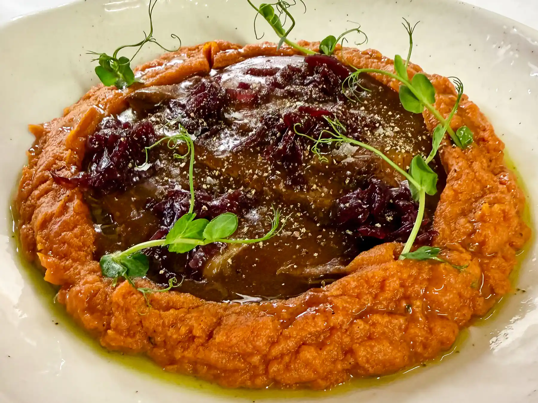 Auf einem hellen, tiefen Teller liegt eine großflächige, glänzende Scheibe dunkelbraunen Fleisches, von Sauce überzogen. Sie ruht in einem breiten Ring aus samtig-orangem Püree, das den Teller fast vollständig umschließt. Dunkelrote, weich wirkende Tupfer und feine grüne Kräuter setzen Kontraste, während ein leichter Ölglanz und feiner Abrieb dem Gericht eine warme, mediterrane Eleganz verleihen.