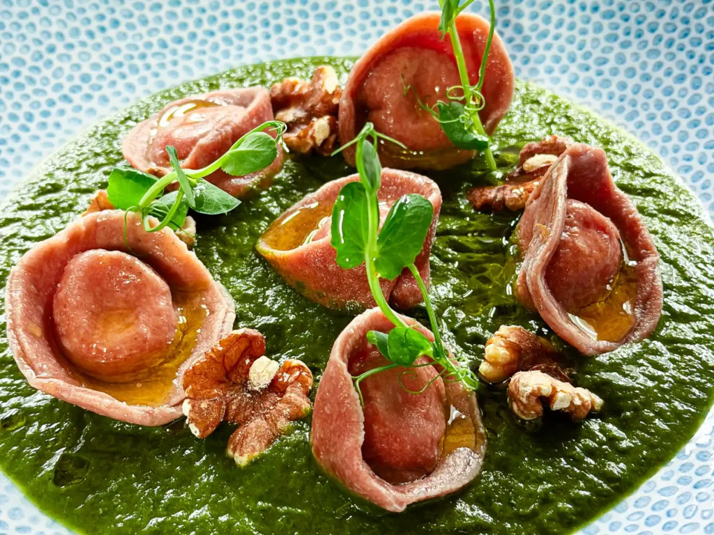 Auf einem hellblauen, fein gemusterten Teller liegen rosafarbene Tortelli kreisförmig auf einem satten, grünen Püree. Die Pasta glänzt leicht, dazwischen liegen grob gebrochene Nüsse und ein paar Tropfen Öl. Frische grüne Sprossen setzen vertikale Akzente und geben dem Gericht eine klare, mediterrane Farbigkeit.