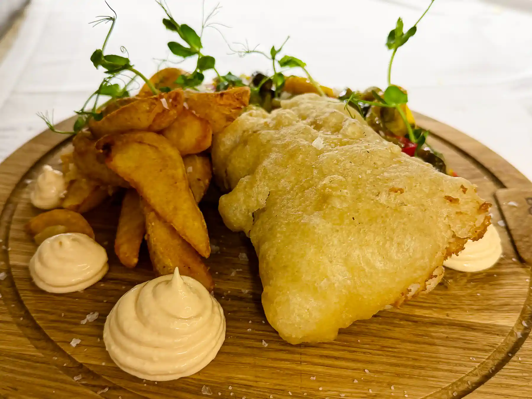 Auf einem runden Holzbrett liegt ein goldgelb ausgebackenes Fischfilet mit luftig-knuspriger Hülle. Daneben stapeln sich rustikal geschnittene, gebräunte Kartoffelspalten. Cremige, hellgelbe Tupfer setzen weiche Akzente, während im Hintergrund eine bunte Mischung aus grünen Kräutern und fein geschnittenem Gemüse Frische und mediterrane Farben ins Bild bringt.