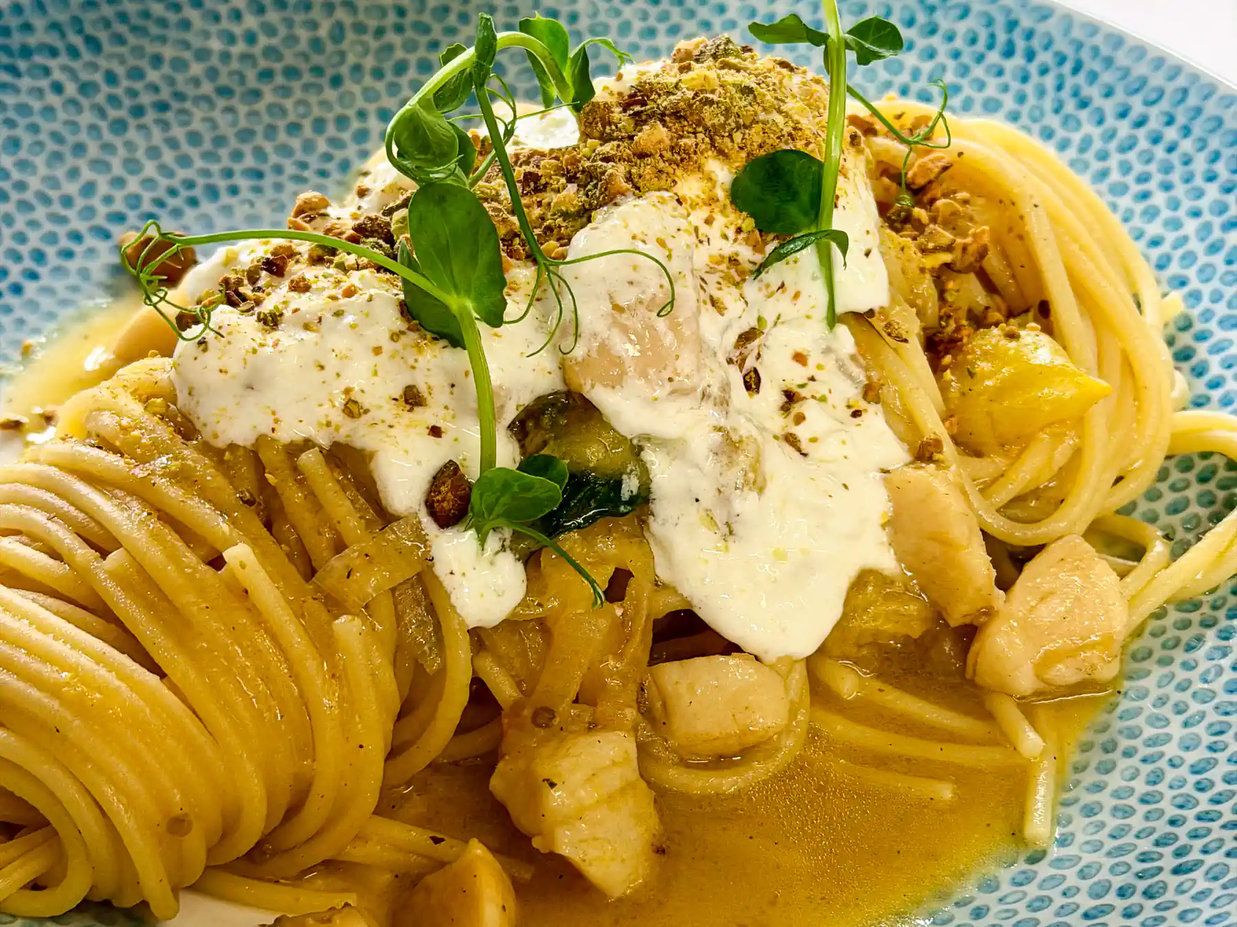 Auf einem hellblauen, fein strukturierten Teller liegt ein großzügig aufgedrehter Nest aus goldgelben Spaghettoni. Die Pasta glänzt von einer warmen, gelblichen Sauce und wird von cremigen, weißen Stracciatella-Flächen überzogen. Dazwischen setzen zart gebräunte, helle Stücke weiche Akzente. Fein gehackte, grünliche Krümel und frische Sprossen bringen Struktur, Höhe und einen frischen, mediterranen Farbkontrast ins Gesamtbild.